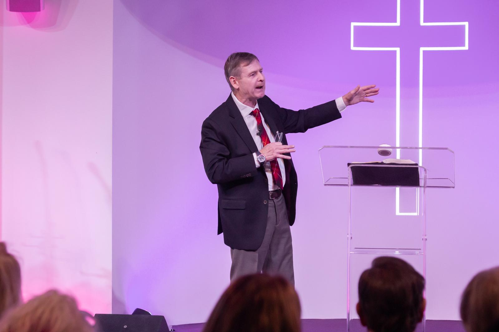 Pastor Rob Andrews preaching before the neon cross, mid-gesture