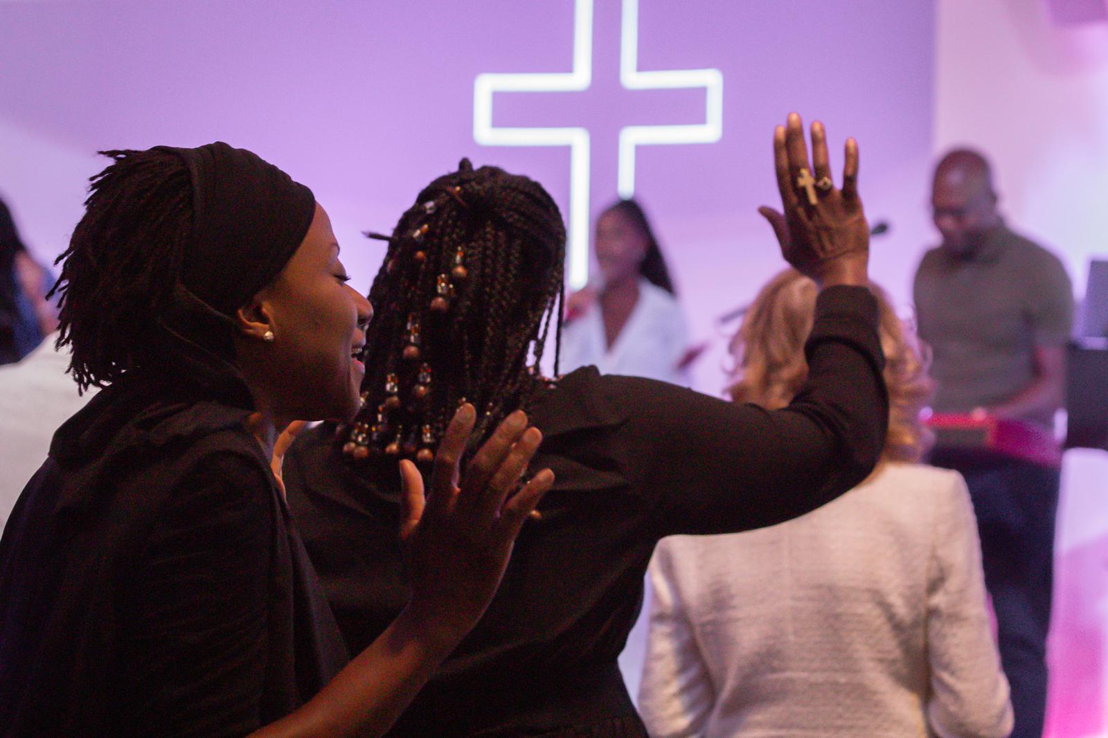 Two women in worship before the neon cross, eyes closed and hand raised
